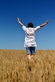 Young woman standing jumping and running  on a wheat field with blue sky in  background at summer day representing healthy life and agriculture concept