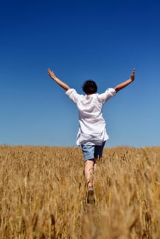 Young woman standing jumping and running  on a wheat field with blue sky in  background at summer day representing healthy life and agriculture concept