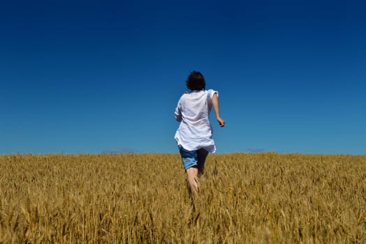 Young woman standing jumping and running  on a wheat field with blue sky in  background at summer day representing healthy life and agriculture concept