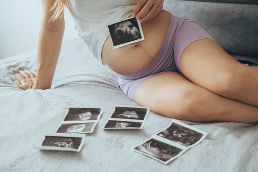 Young pregnant caucasian woman, future mother, sitting on comfortable bed and looking and showing ultrasound x ray photos of her baby in tummy.