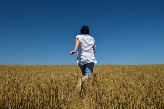 Young woman standing jumping and running  on a wheat field with blue sky the background at summer day representing healthy life and agriculture concept