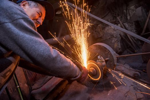 the blacksmith polishing metal products making sparks while using a grinding machine in his traditional workshop