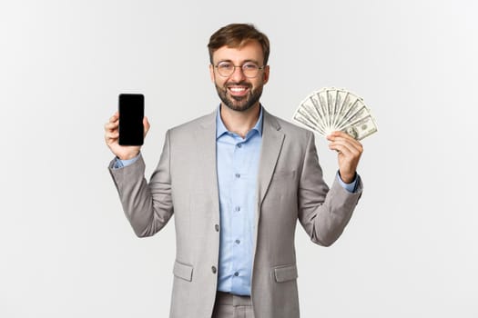 Successful businessman in glasses and grey suit, showing smartphone screen and money, smiling pleased, standing over white background.