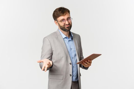 Portrait of male boss with beard, wearing gray suit and glasses, judging bad work, holding digital tablet and looking disappointed, standing over white background.