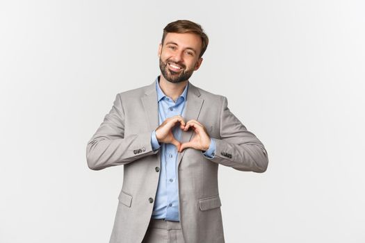 Portrait of handsome smiling bearded man in business suit, showing heart sign, express love or sympathy, standing over white background.