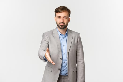 Portrait of confident businessman with beard, wearing grey suit, extending hand for handshake, greeting you, introduce himself, standing over white background.