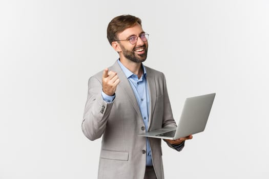Portrait of happy and pleased businessman in grey suit and glasses, working with laptop and pointing finger at camera, praising good idea, standing over white background.