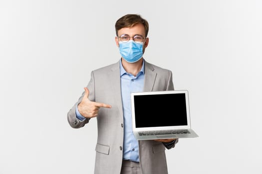 Concept of work, covid-19 and social distancing. Portrait of businessman in medical mask, pointing finger at laptop screen, showing presentation, standing over white background.