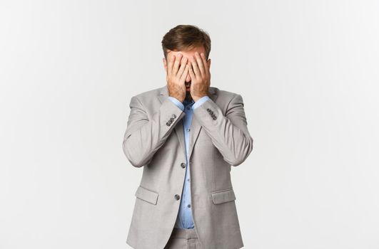 Portrait of distressed and exhausted businessman in grey suit, hiding his face behind hands, standing over white background.