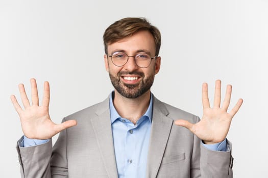 Close-up of handsome bearded man in glasses and gray suit, showing number ten and smiling, standing over white background.