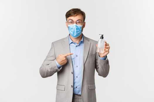 Concept of work, covid-19 and social distancing. Image of male employer in gray suit and medical mask, pointing at hand sanitizer or antiseptic, standing over white background.