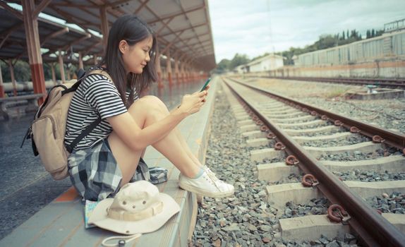 Concept of travel and tourism. Young Asian female tourist uses a mobile phone while waiting for a train at a train station.