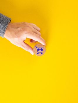 Male hand placing a blue shopping cart icon on yellow background. Shopping or online e-commerce concept.