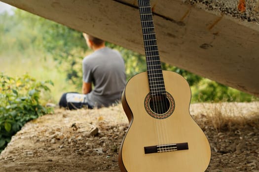 Acoustic guitar, six strings. Boy is sitting on raised platform. Summer day.