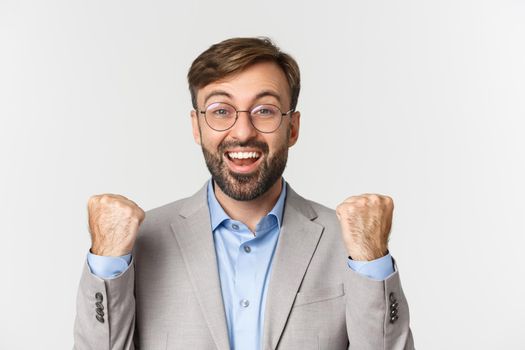 Close-up of successful businessman in glasses and gray suit, rejoicing, saying yes with happy face and making fist pump, achieve goal, standing over white background.