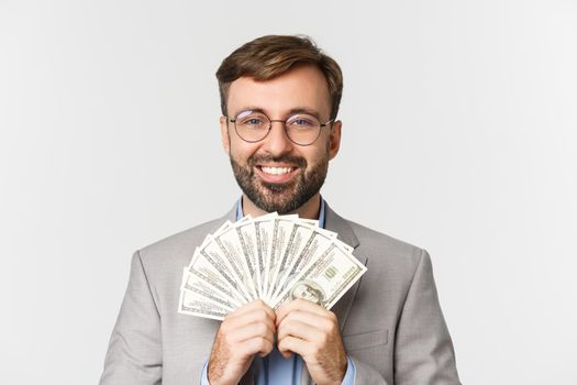 Close-up of rich and successful businessman, wearing gray suit and glasses, showing cash and smiling happy, standing over white background.