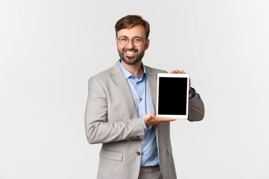 Image of successful and confident male entrepreneur in grey suit and glasses, demonstrating something on digital tablet screen and smiling, standing over white background.