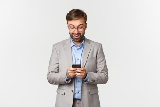 Portrait of handsome businessman with beard, wearing glasses and grey suit, looking surprised at mobile phone, reading something online, standing over white background.