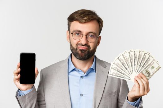 Close-up of sad rich businessman holding money and showing smartphone screen, standing unamused in gray suit and glasses.