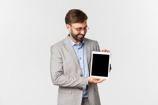 Portrait of happy and satisfied businessman in gray suit and glasses, looking and showing digital tablet screen, smiling proud, standing over white background.