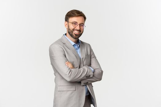 Portrait of confident businessman with beard, wearing grey suit and glasses, cross arms on chest and smiling self-assured, standing over white background.