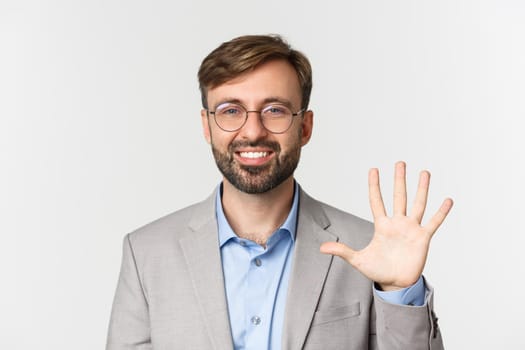 Close-up of handsome bearded man in glasses and gray suit, showing number five and smiling, standing over white background.