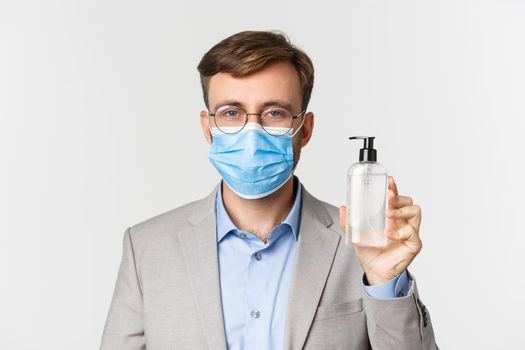 Concept of work, covid-19 and social distancing. Close-up of male boss in gray suit and medical mask, showing hand sanitizer or antiseptic, standing over white background.