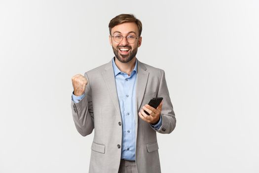 Portrait of cheerful businessman with beard, wearing glasses and grey suit, achieve daily goal in app, making fist pump and holding smartphone, standing happy over white background.