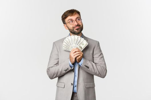 Image of happy businessman with beard, wearing grey suit and glasses, holding money and thinking about shopping or investing, standing over white background.