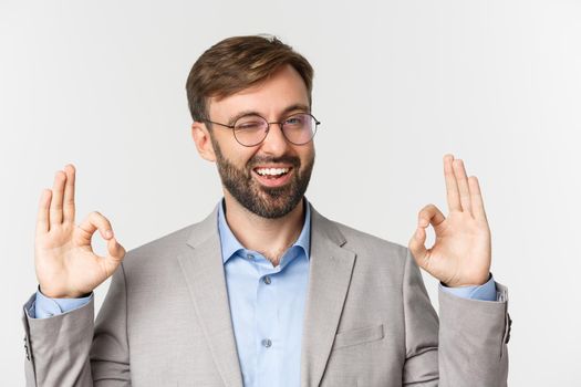 Close-up of impressed boss in gray suit and glasses, showing okay sign and smiling, give approval, praise good job, standing over white background.