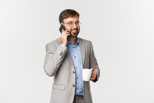 Image of successful businessman in grey suit and glasses, drinking coffee and talking on phone with pleased smile, standing over white background.