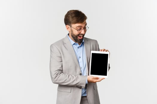 Portrait of excited businessman with beard, wearing glasses and gray suit, demonstrating something on digital tablet screen, looking amazed, standing over white background.
