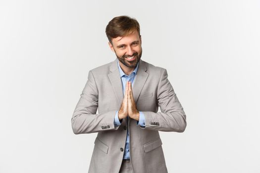 Portrait of happy bearded businessman in grey suit, thanking for something, holding hands pressed together and bowing with grateful expression, standing over white background.