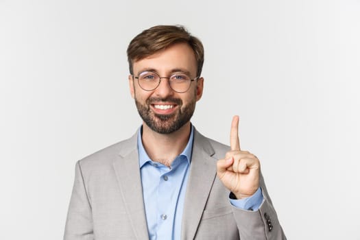 Close-up of handsome bearded man in glasses and gray suit, showing number one and smiling, standing over white background.