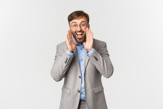 Portrait of happy businessman in grey suit and glasses, making announcement, shouting loud and searching for employees, standing over white background.