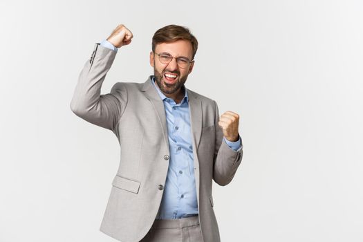 Portrait of successful bearded businessman in grey suit, achieve goal and triumphing, making fist pump gesture and saying yes with confident look, standing over white background.