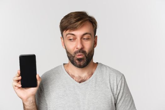 Close-up of excited handsome man in gray t-shirt, showing mobile phone screen, recommending app or shopping site, standing over white background.