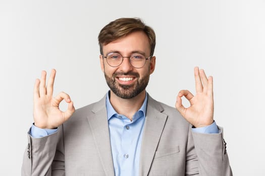 Close-up of handsome confident businessman, wearing glasses and gray shirt, showing okay sign and smiling, approve something good, standing over white background.