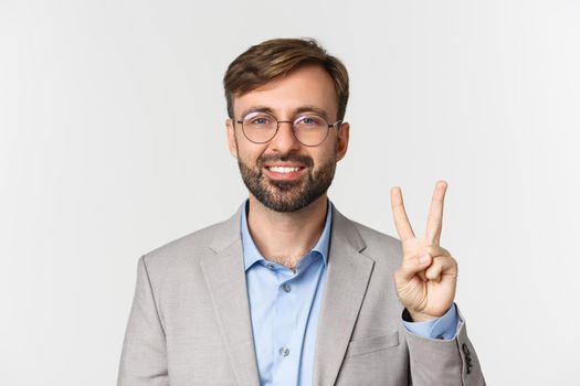 Close-up of handsome bearded man in glasses and gray suit, showing number two and smiling, standing over white background.