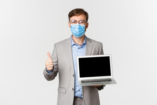 Concept of work, covid-19 and social distancing. Handsome confident businessman in medical mask, showing laptop screen and thumbs-up in approval, standing over white background.