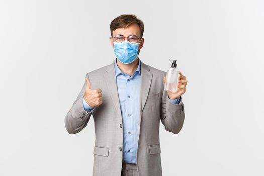 Concept of work, covid-19 and social distancing. Image of handsome businessman in gray suit and medical mask advice using hand sanitizer, showing thumbs-up in approval.