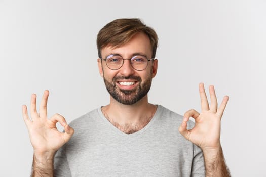 Close-up of cheerful guy with beard, wearing glasses and casual t-shirt, smiling and showing okay sign in approval, recommend product, standing over white background.