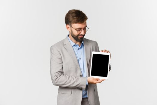 Portrait of smiling bearded businessman in glasses and gray suit, looking kindly at digital tablet screen, satisfied with something, standing over white background.