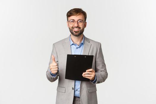 Portrait of satisfied businessman in gray suit and glasses, holding clipboard and showing thumbs-up in approval, approve something good,s tanding over white background.