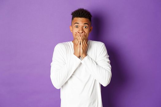 Image of shocked african-american hipster guy in white sweatshirt, gasping and looking startled, cover mouth with hands, standing over purple background.