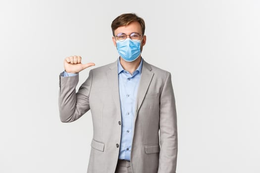 Concept of work, covid-19 and social distancing. Happy businessman in glasses, grey suit and medical mask pointing at himself, showing example to employees, standing over white background.