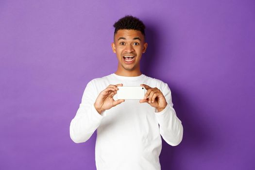 Young excited african-american guy taking pictures on smartphone, holding mobile phone and recording video with amazed face expression, standing over purple background.