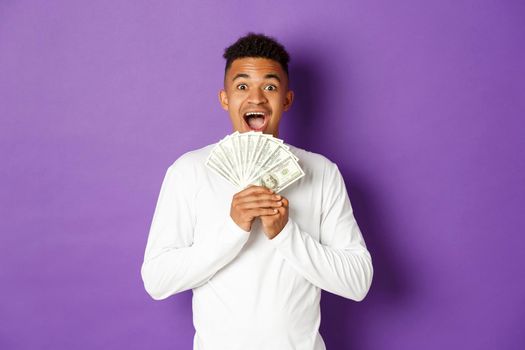 Excited african-american guy in white sweatshirt, holding money and looking amazed, standing over purple background.