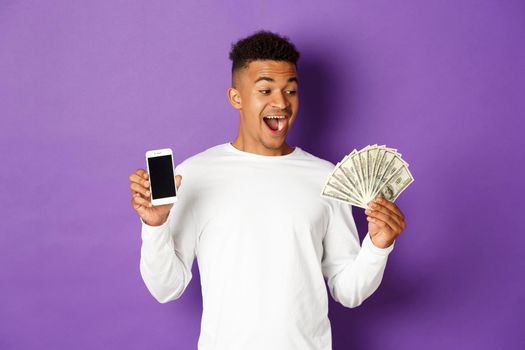 Portrait of modern african-american guy, showing money and mobile phone screen, smiling amazed, standing over purple background.