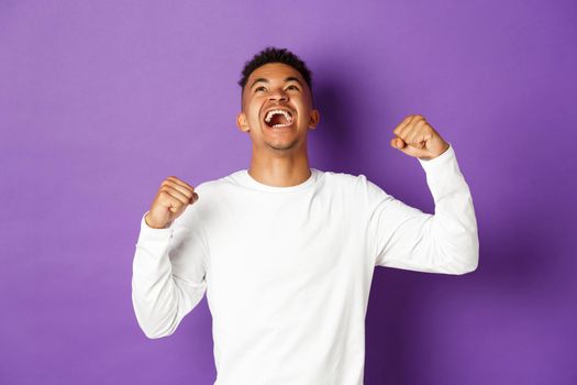 Image of happy and relieved african-american man, celebrating victory, looking up and making fist pump gesture, shouting yes with joyful expression, standing over purple background.